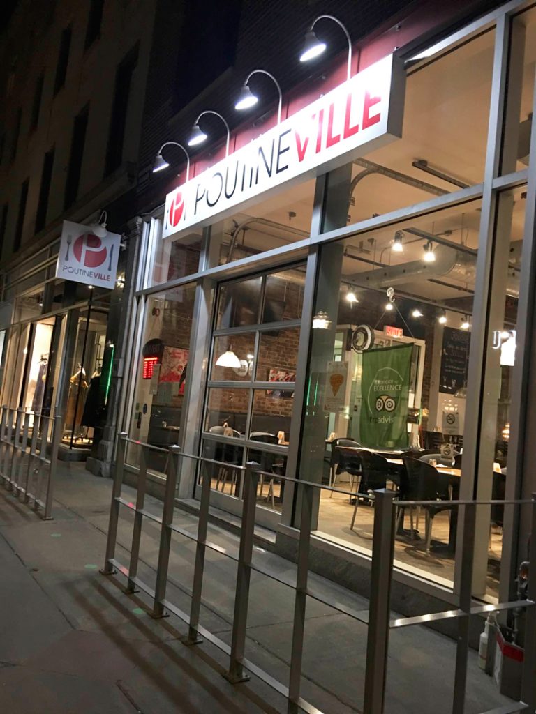 Poutineville, vegan food in Qu&eacute;bec City. Exterior of Poutineville restaurant in Quebec City at night, with modern glass windows and interior lights visible through the storefront. The front entrance of Poutineville glows under bright lights on a quiet street in Quebec City. Tazim found it easy and safe to visit solo in the evening, with welcoming staff and a casual vibe.