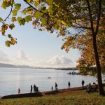 A tranquil waterfront scene with people walking along the shore, framed by colorful autumn leaves and a distant view of the Lion&rsquo;s Gate Bridge. This captures the essence of enjoying nature and scenic views during fall in Vancouver.