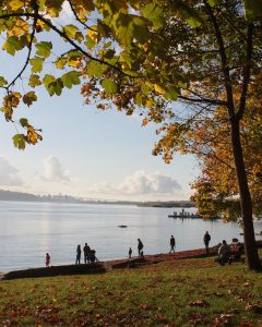 A tranquil waterfront scene with people walking along the shore, framed by colorful autumn leaves and a distant view of the Lion&rsquo;s Gate Bridge. This captures the essence of enjoying nature and scenic views during fall in Vancouver.