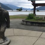 Sasquatch statue standing near a calm lake with distant mountains framed by an overcast sky. This quirky attraction adds a touch of fun to springtime exploration around Vancouver, offering a unique photo opportunity amidst natural beauty.