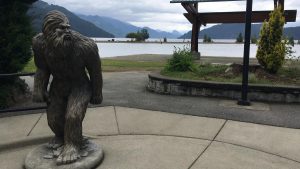 Sasquatch statue standing near a calm lake with distant mountains framed by an overcast sky. This quirky attraction adds a touch of fun to springtime exploration around Vancouver, offering a unique photo opportunity amidst natural beauty.