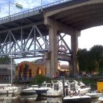 Boats moored at Granville Island under the bridge, with the vibrant Public Market entrance visible in the background. This image emphasizes Granville Island's lively waterfront and market scene, ideal for a spring outing.