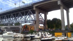 Boats moored at Granville Island under the bridge, with the vibrant Public Market entrance visible in the background. This image emphasizes Granville Island's lively waterfront and market scene, ideal for a spring outing.