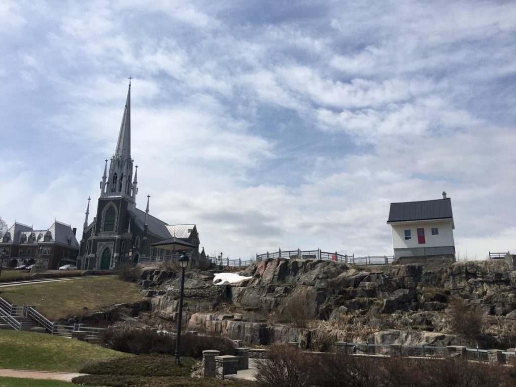 A stunning Gothic-style church with a towering spire sits atop a hill in Saguenay, Quebec. Nearby, a tiny, modern house perches on a rocky outcrop, creating a striking contrast between historical and contemporary architecture.