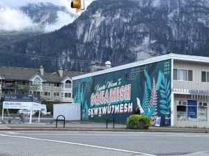 This image showcases a colorful mural on the side of a building, reading "Kw'achn&aacute;m Welcome to Squamish Skwxw&uacute;7mesh" in bold lettering. The mural features vibrant designs of plants and is labeled "Sea and Sky" in the top corner. Behind the building, steep mountain cliffs rise into the sky, partially covered by mist or clouds.
