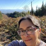 Tazim wearing glasses and a light purple t-shirt takes a selfie while hiking in a scenic mountain area. Behind her, lush greenery, power lines, and distant mountain peaks stretch under a clear blue sky.