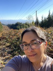 Tazim wearing glasses and a light purple t-shirt takes a selfie while hiking in a scenic mountain area. Behind her, lush greenery, power lines, and distant mountain peaks stretch under a clear blue sky.