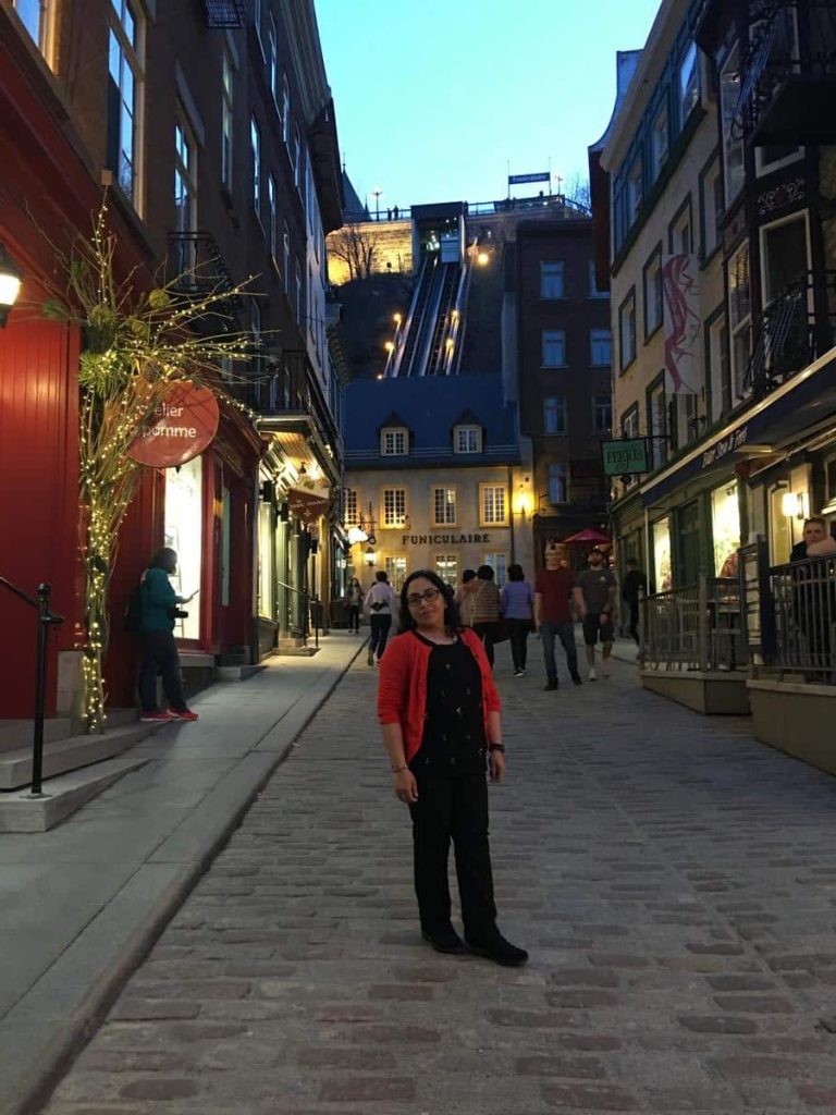 Tazim Quartier Petit Champlain. Tourist standing on Rue du Petit-Champlain in Old Quebec, with the funicular visible in the background and shops lining both sides of the cobblestone street.