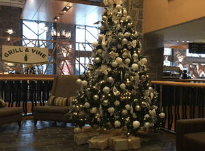 A large, decorated Christmas tree in a hotel lobby, adorned with white and gold ornaments, lights, and wrapped presents at the base. The space includes cozy chairs, with the "Grill & Vine" restaurant sign in the background.