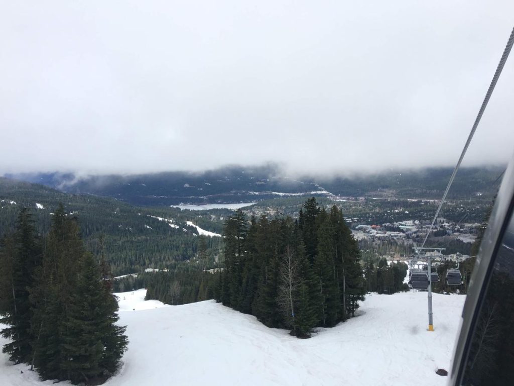 A view from a gondola overlooking a snowy mountainside with evergreen trees. The gondola cable stretches towards a foggy sky, with a small town and lake barely visible through the mist in the valley below.