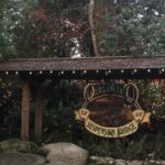 The entrance to the Capilano Suspension Bridge, featuring a rustic wooden sign with lights and surrounded by festive red and white holiday decorations in a lush forest.