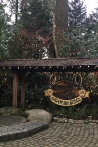 The entrance to the Capilano Suspension Bridge, featuring a rustic wooden sign with lights and surrounded by festive red and white holiday decorations in a lush forest.