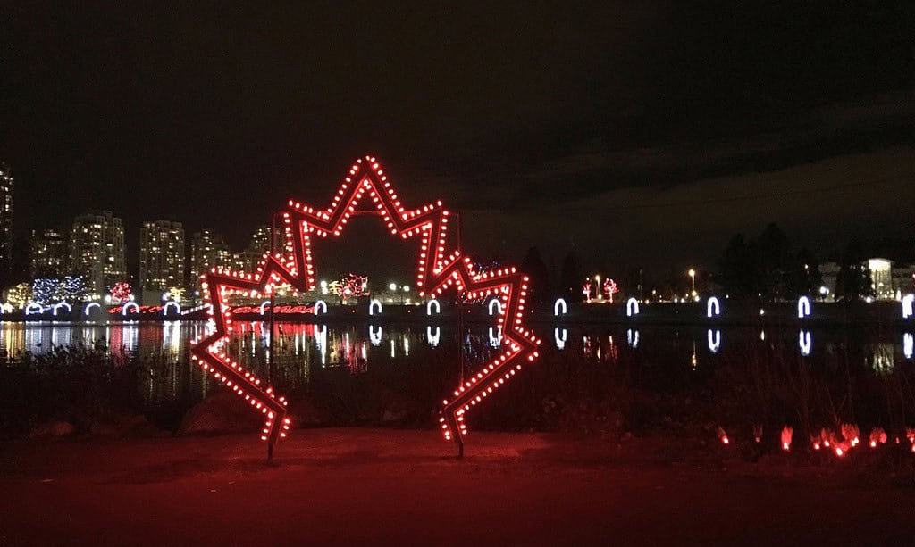 A serene night view of a lake bordered by arches wrapped in white lights, with brightly lit trees reflecting in the water. A lit-up maple leaf.