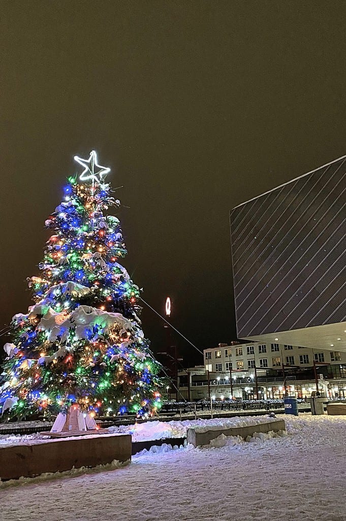 A festive Christmas tree decorated with colorful lights and a glowing star topper, surrounded by a snowy plaza at night. The scene includes modern buildings and snow-covered ground, enhancing the holiday ambiance.