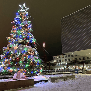 A festive Christmas tree decorated with colorful lights and a glowing star topper, surrounded by a snowy plaza at night. The scene includes modern buildings and snow-covered ground, enhancing the holiday ambiance.