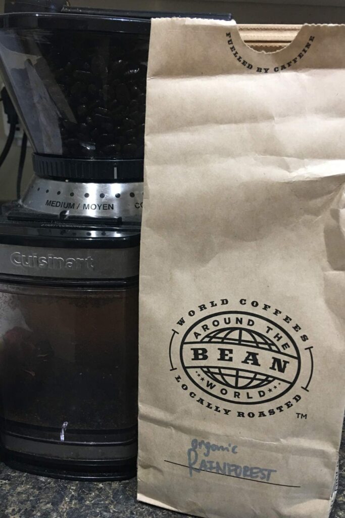 A brown paper bag of organic "Rainforest" locally roasted coffee next to a grinder filled with dark coffee beans. It highlights the availability of premium coffee beans in North Vancouver.