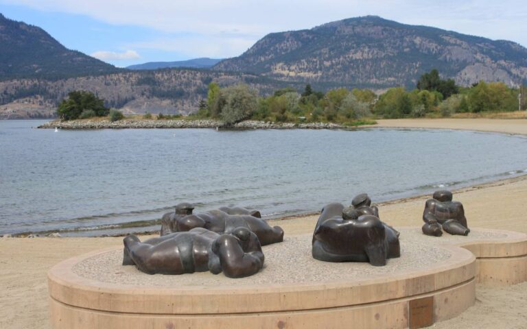 Sculptures by Okanagan Lake Bronze sculptures of reclining figures on a sandy beach with Lake Okanagan and mountains in the distance. This illustrates Kelowna&rsquo;s artistic side and its relaxing lakeside ambiance.