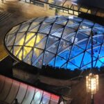 An aerial view of the illuminated glass dome covering the Robson Square ice skating rink at night, with a mix of blue and golden lights shining through, surrounded by urban architecture.