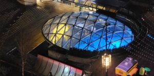 An aerial view of the illuminated glass dome covering the Robson Square ice skating rink at night, with a mix of blue and golden lights shining through, surrounded by urban architecture.