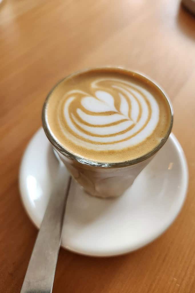 A perfectly crafted latte with intricate heart-shaped foam art sits on a white saucer on a wooden table. North Vancouver's coffee culture shines through in the artistry and quality of drinks like this.