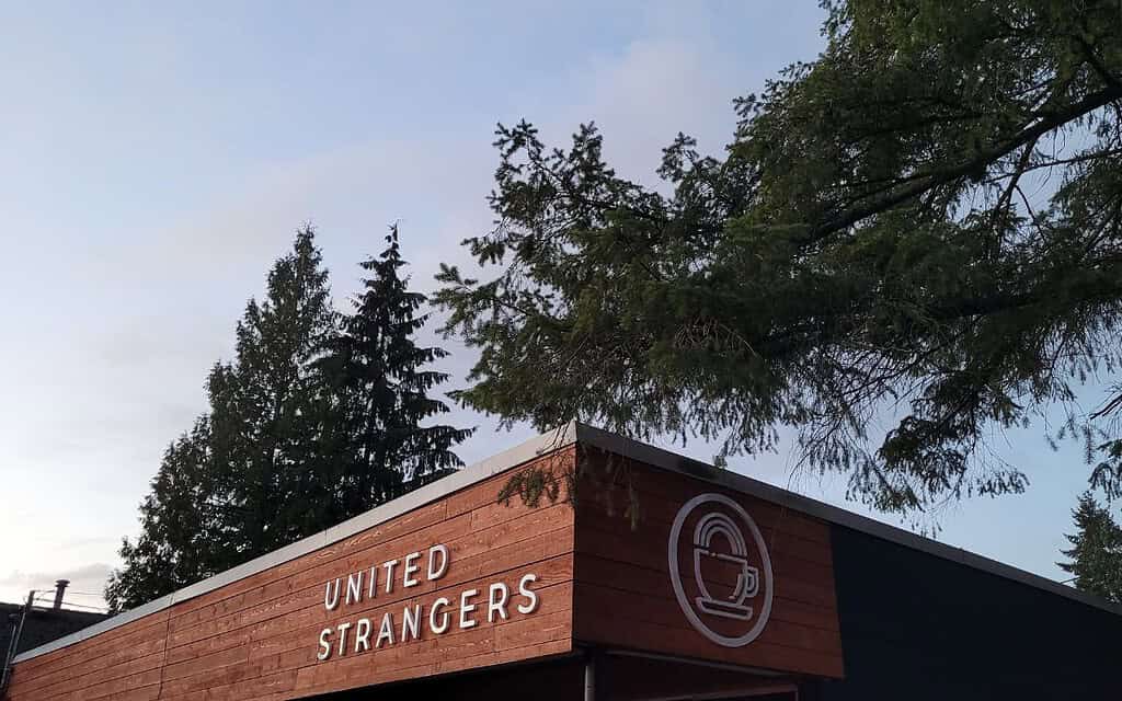 A cozy cafe with a wooden exterior and modern logo is surrounded by tall evergreen trees under a light blue sky. United Strangers is a must-visit coffee spot in North Vancouver, offering a perfect blend of nature and great coffee.