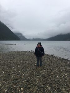 Tazim Damji standing on the rocky shore with mountains and dark clouds in the background.