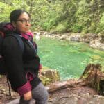 Tazim Damji stands by the vibrant turquoise waters of the Capilano River, surrounded by towering trees, making it a picture-perfect spring hiking destination.