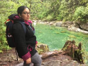 Tazim Damji stands by the vibrant turquoise waters of the Capilano River, surrounded by towering trees, making it a picture-perfect spring hiking destination.