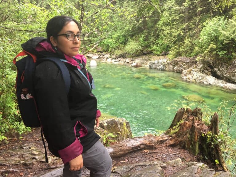 Tazim Damji stands by the vibrant turquoise waters of the Capilano River, surrounded by towering trees, making it a picture-perfect spring hiking destination.