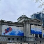 The Vancouver Art Gallery, featuring grand neoclassical architecture and banners for a Firelei B&aacute;ez exhibition. The banners display vibrant artwork, adding a splash of color to the gray, overcast day.