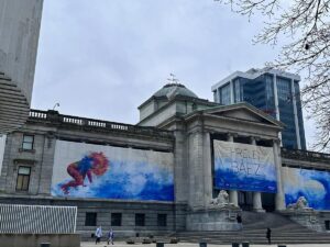 The Vancouver Art Gallery, featuring grand neoclassical architecture and banners for a Firelei B&aacute;ez exhibition. The banners display vibrant artwork, adding a splash of color to the gray, overcast day.