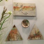 A collection of hand-painted ceramic art pieces displayed on a gallery wall, including designs featuring birds and horses.