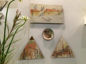 A collection of hand-painted ceramic art pieces displayed on a gallery wall, including designs featuring birds and horses.