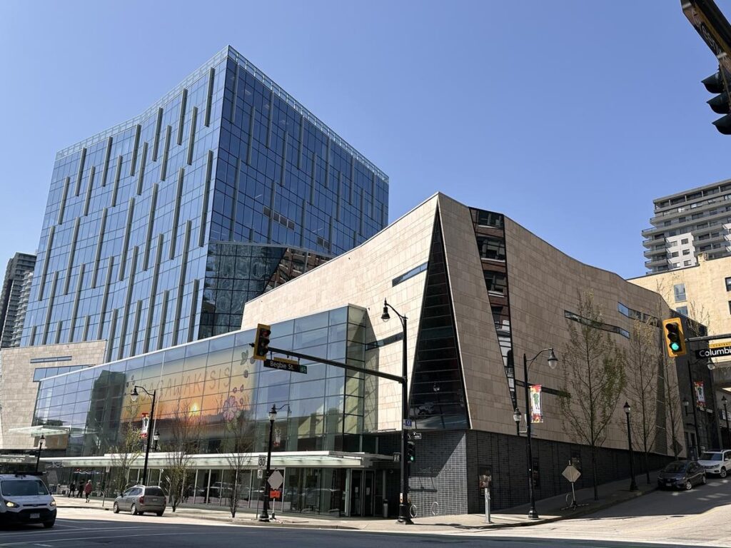 A sleek modern building with glass panels and beige stone walls, standing at a busy intersection under a clear blue sky. The Anvil Centre anchors downtown New Westminster and makes it easy to find so many things to do nearby.