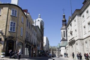 Rue Saint-Louis bustling with people, framed by classic stone buildings and a church steeple. The street is a vital artery in Old Quebec, connecting visitors to landmarks, shops, and eateries.
