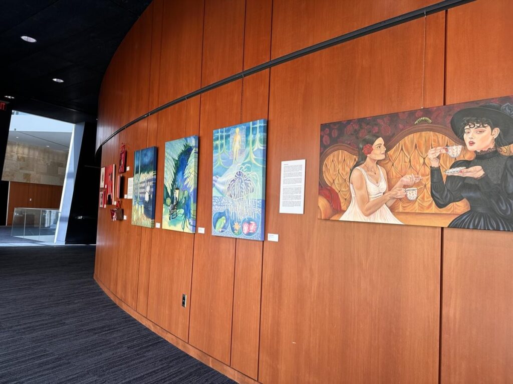 Bright hallway with wooden walls displaying colorful local paintings, including a vibrant scene of two women having tea. Shows off the local art that fills public spaces and adds character to downtown.