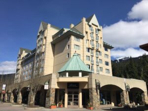 The grand exterior of Fairmont Chateau Whistler features a peaked green roof, arched stone entryways, and alpine-inspired architecture beneath a clear blue sky. This iconic Whistler hotel sits at the base of Blackcomb Mountain with instant village access.