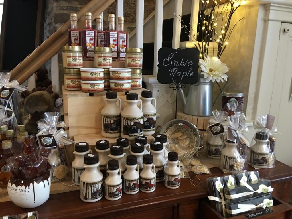 A cozy display of maple syrup products and treats in a shop corner labeled &ldquo;&Eacute;rable Maple,&rdquo; featuring bottles, jars, and candies artfully arranged near soft lighting and rustic decor. The scene highlights Quebec&rsquo;s signature maple goods.