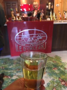 A hand holds a glass of golden cider or wine in front of a red counter labeled &ldquo;&Icirc;le d&rsquo;Orl&eacute;ans,&rdquo; with a woman behind the tasting bar surrounded by bottles. The cozy setting suggests a local Quebec tasting experience.