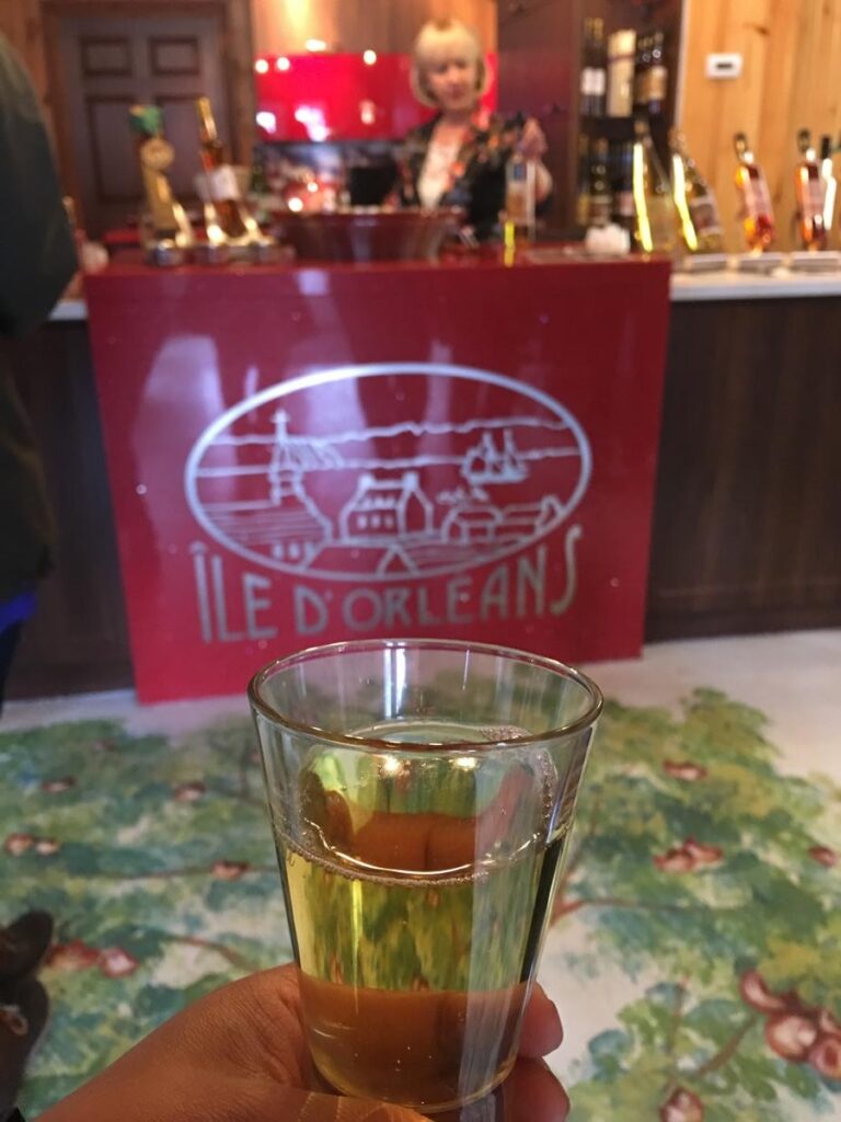 A hand holds a glass of golden cider or wine in front of a red counter labeled &ldquo;&Icirc;le d&rsquo;Orl&eacute;ans,&rdquo; with a woman behind the tasting bar surrounded by bottles. The cozy setting suggests a local Quebec tasting experience.