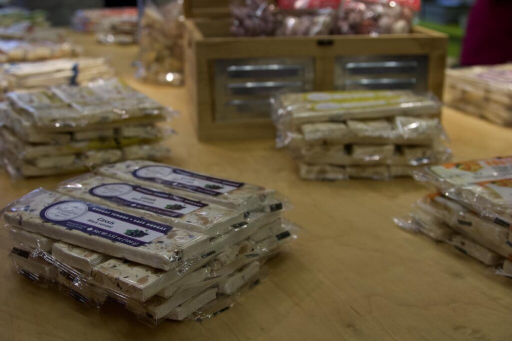 Stacks of packaged nougat bars in flavors like blackcurrant and citrus are displayed on a wooden table in a specialty shop. The packaging is labeled &ldquo;Nougat tendre - soft nougat,&rdquo; highlighting this sweet Quebec treat.