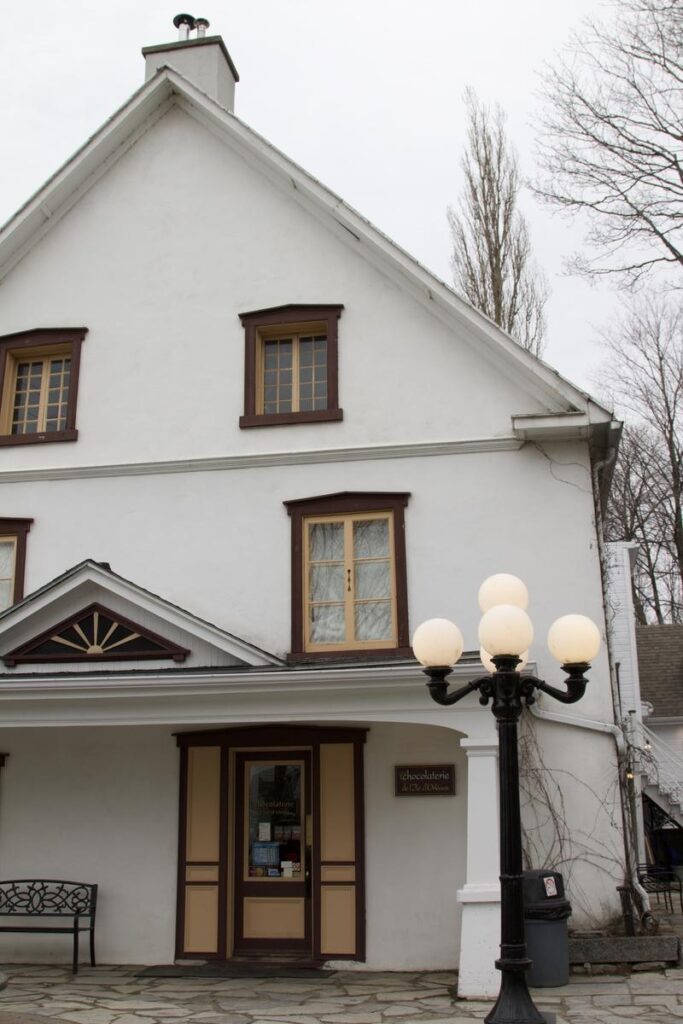 The front of a white historic-style building sits under a cloudy sky, complete with a lamp post and a stone walkway. The welcoming facade suggests a local artisan chocolate shop as one of the best things to do on &Icirc;le d&rsquo;Orl&eacute;ans.
