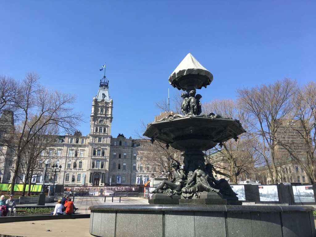 Foreground fountain with sculpted figures in front of Quebec&rsquo;s Parliament Building, seen under a bright blue sky with visitors sitting nearby. The tall central tower with its clock and Quebec flag rises behind the ornate fountain.