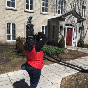 Tazim takes a photo of a bronze statue in front of a building with a bright red door labeled "Monast&egrave;re." The walkway and landscaping suggest a peaceful courtyard setting.