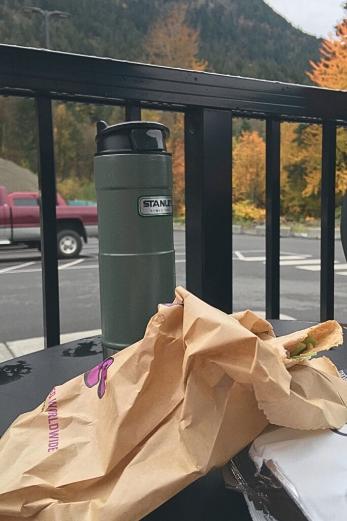 A Stanley bottle and wrapped sandwich sit on an outdoor table with fall trees and mountains in the background. A simple road trip lunch stop near Manning Park is exactly the kind of low-effort planning that makes the day feel easier.