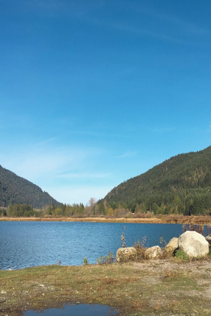 A blue mountain lake sits below forested hills in Manning Park, with large rocks and dry grass along the shore. This is the kind of quiet lake view that makes Manning Park feel worth the drive from Vancouver.