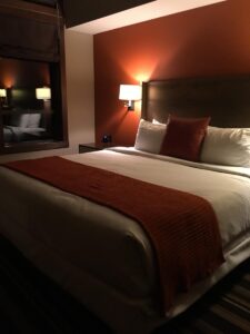 A spacious king room at Evolution Creekside Hotel in Whistler shows the bed centered against a rust-orange accent wall, with bedside lamps and a soft striped carpet. Tucked away from the main village, it&rsquo;s a stylish, peaceful retreat with a modern alpine touch.