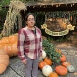 Woman wearing a red plaid button-up and grey pants poses by a pumpkin display at Capilano Suspension Bridge&mdash;casual outfit for fall in Vancouver, Canada.
