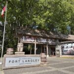A rustic wooden building stands beneath tall green trees, with a Canadian flag in front and signage reading &ldquo;Fort Langley.&rdquo; Perfect for a day trip from Vancouver, the visitor centre at Fort Langley National Historic Site welcomes guests with artwork that tells stories of the region&rsquo;s past.