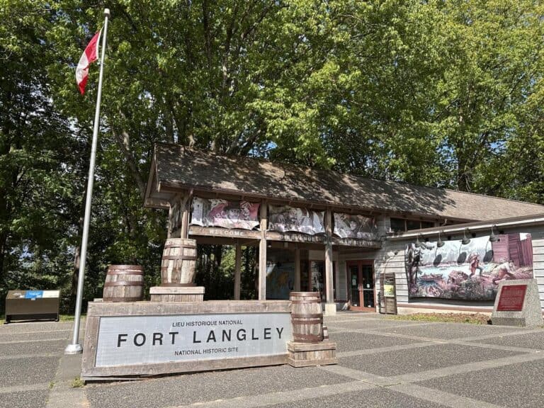 A rustic wooden building stands beneath tall green trees, with a Canadian flag in front and signage reading &ldquo;Fort Langley.&rdquo; Perfect for a day trip from Vancouver, the visitor centre at Fort Langley National Historic Site welcomes guests with artwork that tells stories of the region&rsquo;s past.